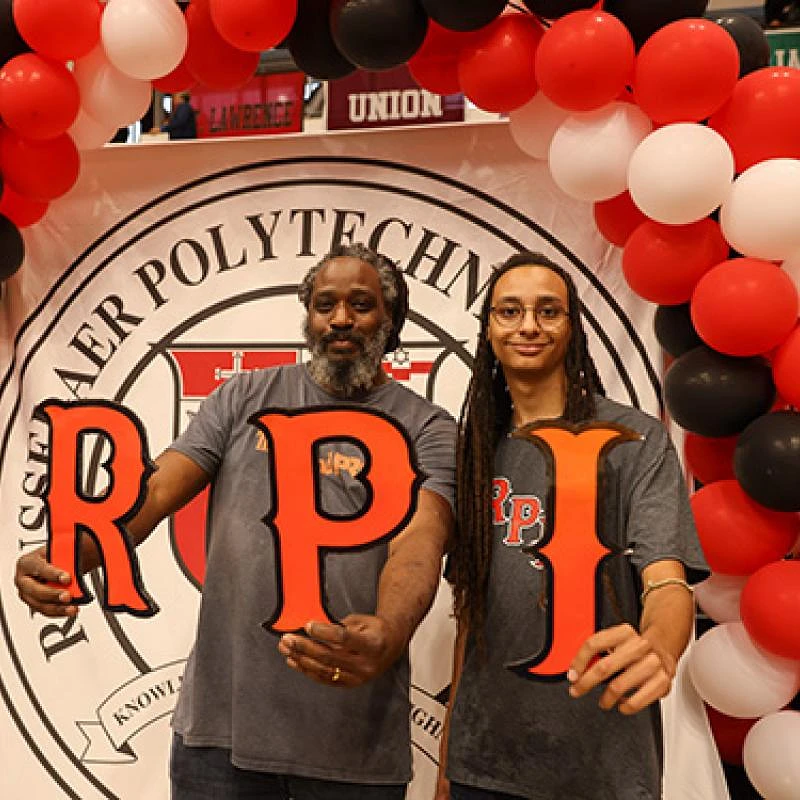 Two people are holding large letters spelling "RPI" in front of a Rensselaer Polytechnic Institute banner, surrounded by red, white, and black balloons.